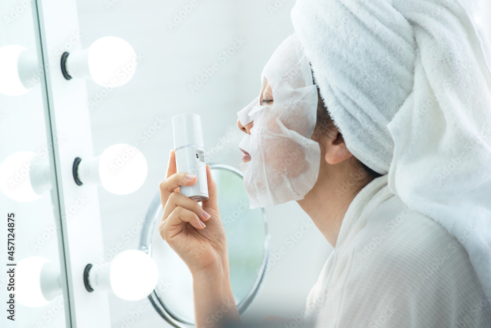 Beautiful girl in bathrobe and turban towel on her head, mask with sheet mask and spray mineral water on the mask in front of the mirror, women's lifestyle Beautiful girl in bathrobe and turban towel on her head, mask with sheet mask and spray mineral water on the mask in front of the mirror, women's lifestyle