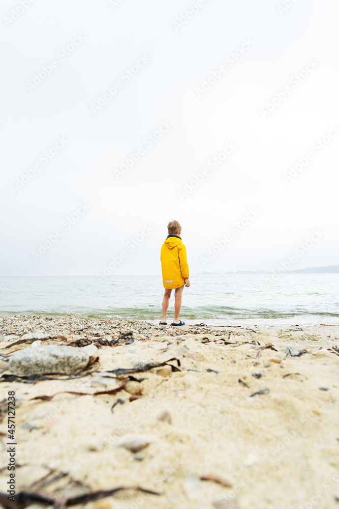 A little boy child in a yellow windbreaker stands on the sandy shore of the ocean all alone.