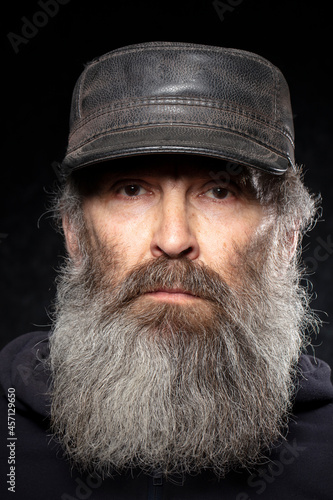 The face of an elderly man with a gray beard in a leather hat on a black background.