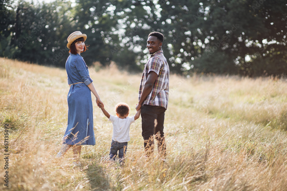 Back view of little african boy holding hands with caring parents while standing on summer nature. Caucasian woman and african man smiling and looking at camera. Family and love concept.
