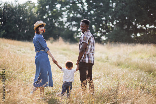 Adorable multi ethnic family of three in casual clothes walking together on summer field. Young and happy parents holding hands with little son and smiling sincerely on camera.