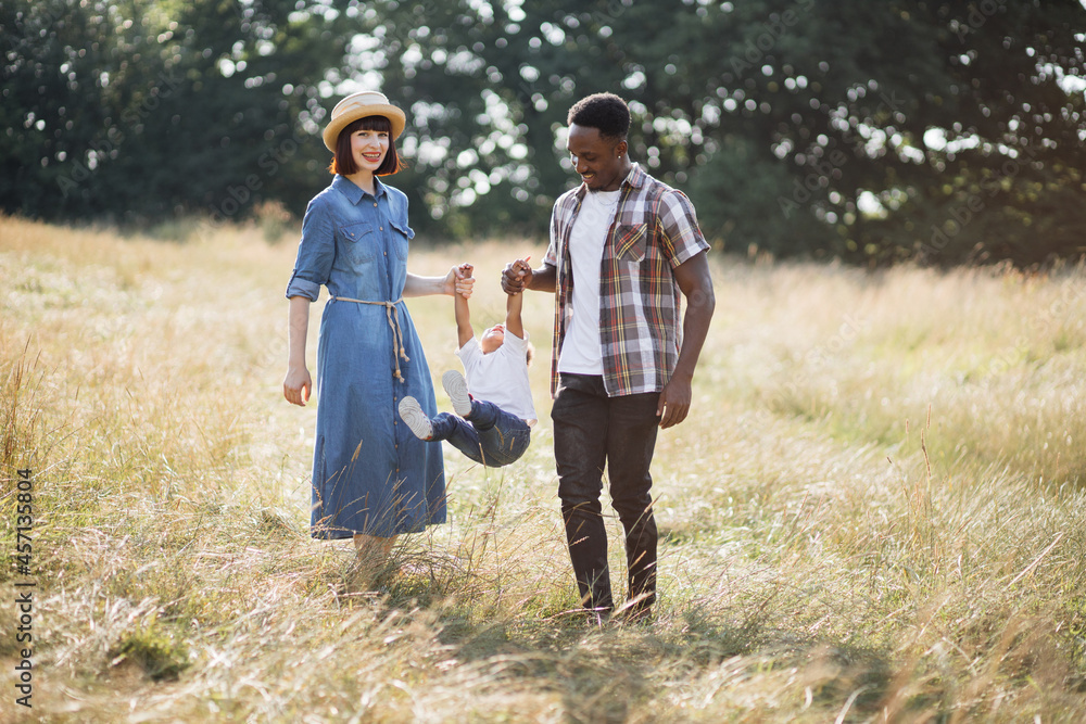 Multiracial young parents raising son for both arms while playing on summer field. Cheerful diverse family in causal outfit spending time together on nature. Multiracial young parents raising son for both arms while playing on summer field. Cheerful diverse family in causal outfit spending time together on nature.