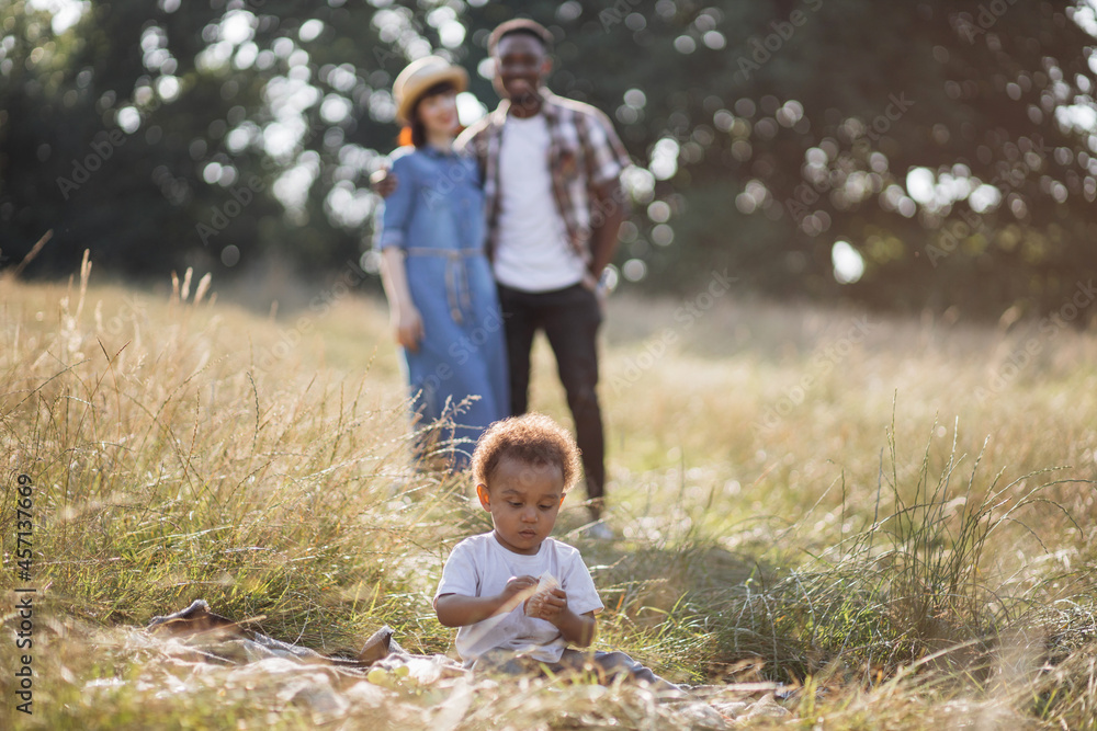 Fototapeta premium Blur background of multicultural parent hugging and smiling on field while their cute son sitting on checkered blanket and playing. Summer picnic of happy family. Enjoyment and relaxation concept.