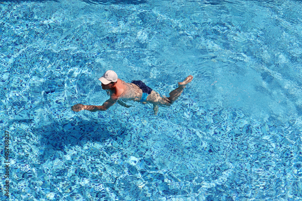 Man in baseball cap swimming in the pool water, top view. Water sports, beach vacation