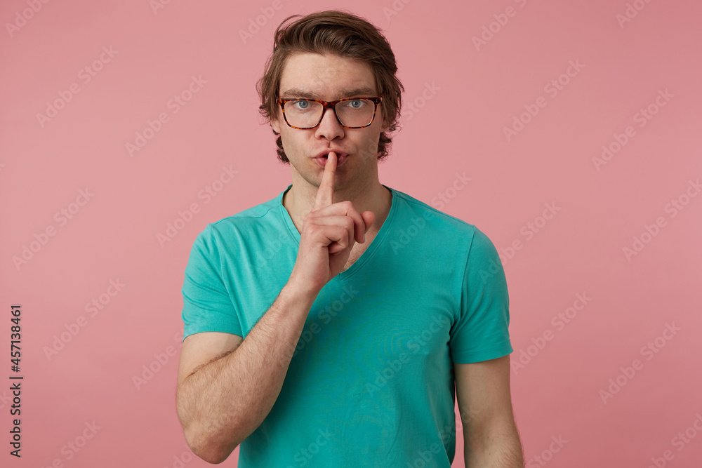 Studio shot of young handsome male, wears glasses and blue t-shirt, looks into camera and show silence gesture with serious facial expression. Isolated over pink background Studio shot of young handsome male, wears glasses and blue t-shirt, looks into camera and show silence gesture with serious facial expression. Isolated over pink background