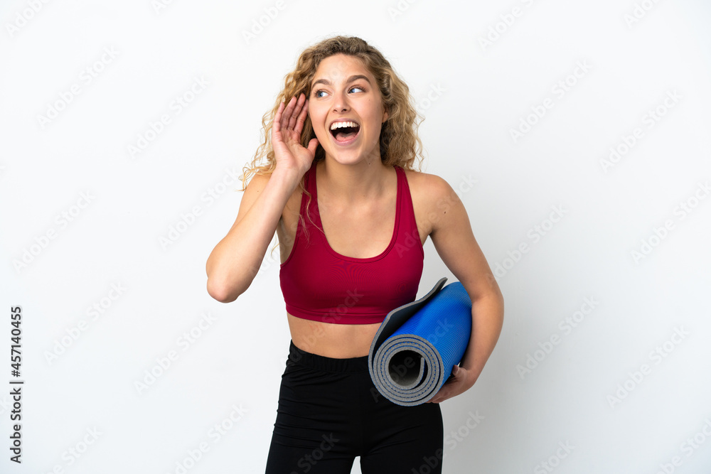 Young sport blonde woman going to yoga classes while holding a mat isolated on white background listening to something by putting hand on the ear Young sport blonde woman going to yoga classes while holding a mat isolated on white background listening to something by putting hand on the ear