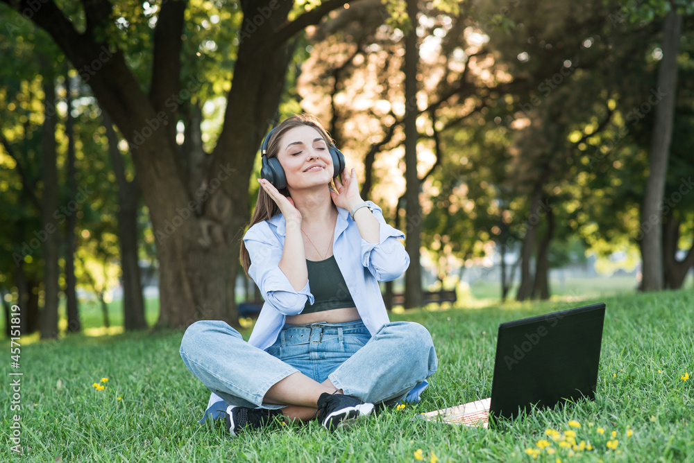 A young and attractive girl in headphones sitting on the grass on the lawn. Using laptop to listening to music with headphones A young and attractive girl in headphones sitting on the grass on the lawn. Using laptop to listening to music with headphones