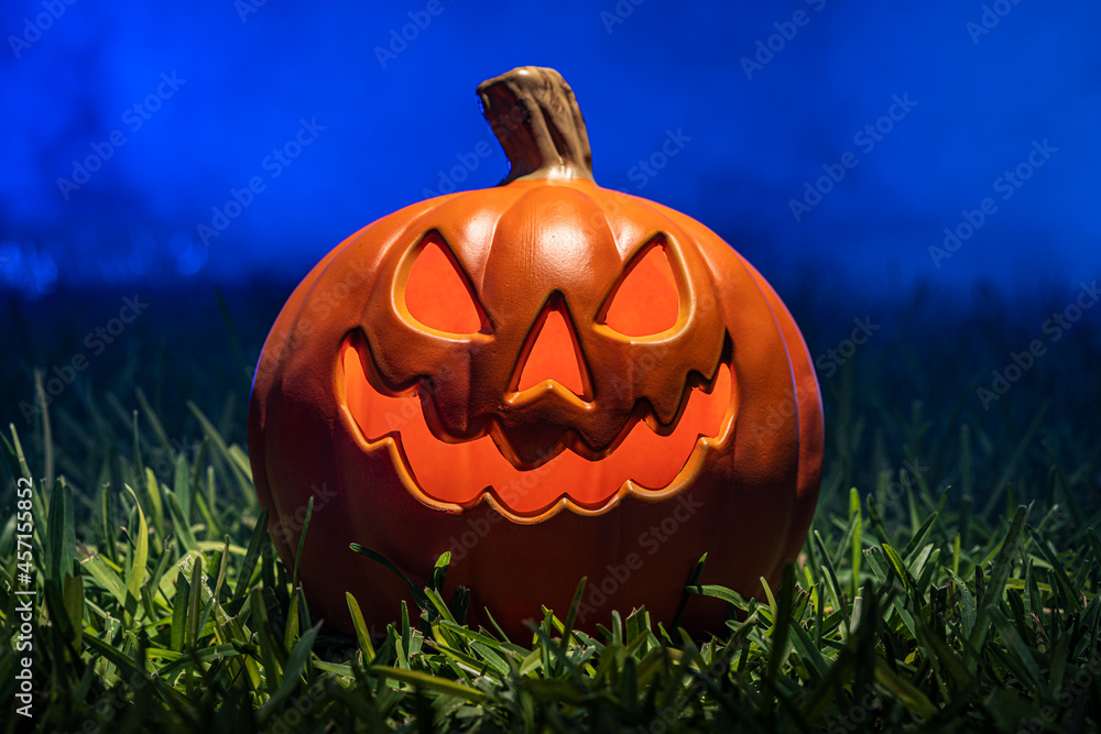 Obraz premium Spooky Scary Orange Pumpkin Creepy Laughing Tooth Smile in Green Grass with Misty Foggy Blue Background