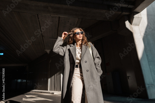 Stylish beautiful young girl with curly hair with sun glasses in a fashionable long coat walks in the city. Female model, urban style and beauty. Sunlight and shadow © alones