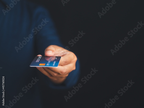 Hand of a young man holding a mockup blue credit card while standing with a black background in the studio. Space for text. Close-up photo. Money and business concept