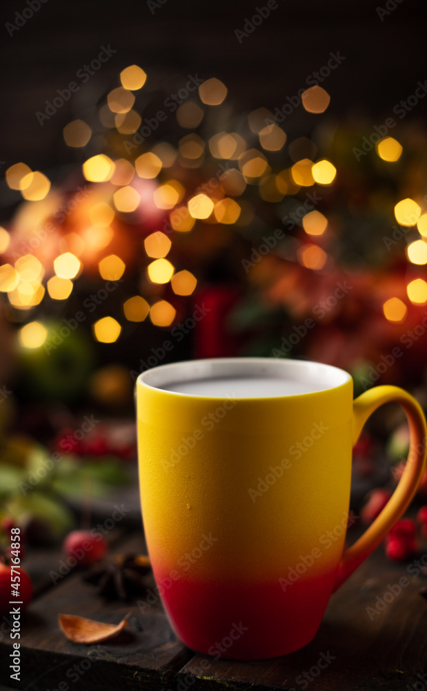 a red and yellow mug with a steaming drink. Fruits and spices are all around on a wooden table. Yellow lights of garlands are burning behind a red and yellow mug with a steaming drink. Fruits and spices are all around on a wooden table. Yellow lights of garlands are burning behind