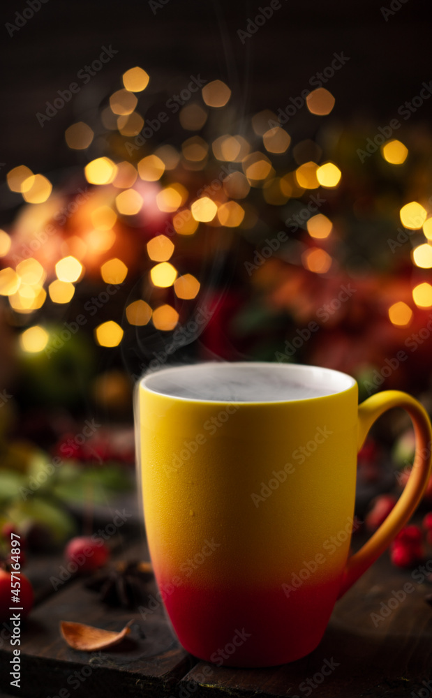 a red and yellow mug with a steaming drink. Fruits and spices are all around on a wooden table. Yellow lights of garlands are burning behind a red and yellow mug with a steaming drink. Fruits and spices are all around on a wooden table. Yellow lights of garlands are burning behind