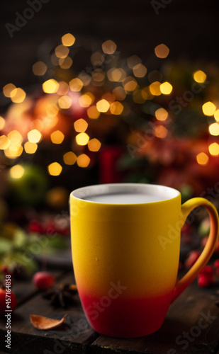a red and yellow mug with a steaming drink. Fruits and spices are all around on a wooden table. Yellow lights of garlands are burning behind