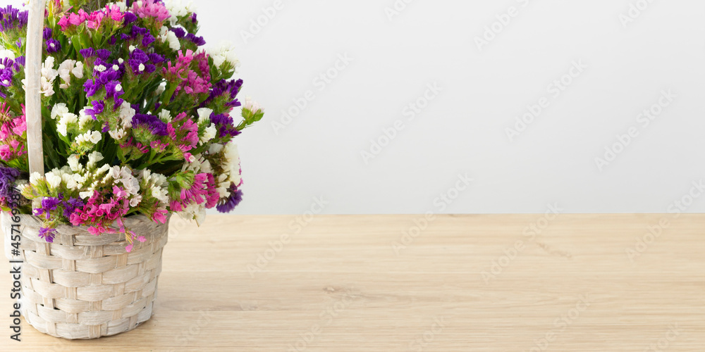A basket with multicolored statice flowers stands on a wooden table top on a light background . The flowers are purple, white and pink . A banner with free space for your business .