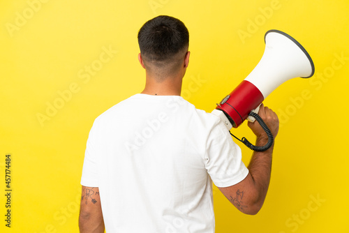Young handsome man over isolated yellow background holding a megaphone and in back position