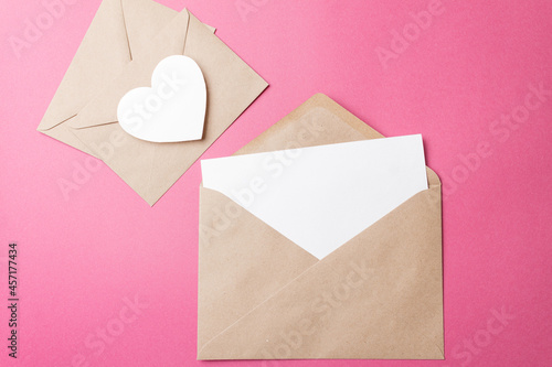 The concept of a romantic love letter for Valentine's Day. A kraft envelope with a blank sheet of paper inside and a white heart on a pink background. An invitation to a wedding. Space for text © Evgenii