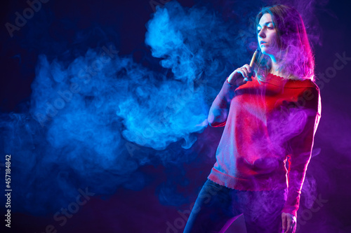 A woman with a vape device in a puff of smoke. The girl smokes a vape on a dark background. The girl in red smokes an e-cigarette. Woman engaged veypingom