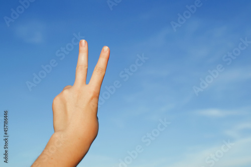 A hand against the background of a blue sky, shows a sign of victory or peace, is shown with the index and middle fingers of the hand pointing upwards in the form of the Latin letter "V", copy space.