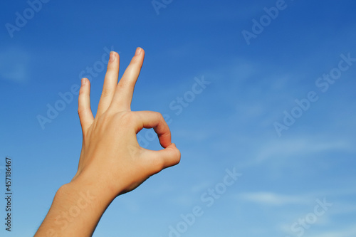 A hand against a blue sky shows an okay gesture, performed by connecting the thumb and forefinger and keeping the other fingers straight or relaxed from the palm, copying space.