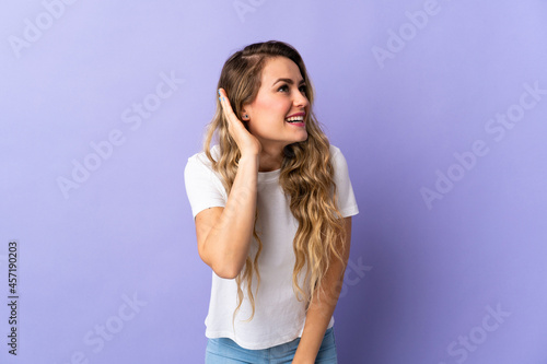 Young Brazilian woman isolated on purple background listening to something by putting hand on the ear