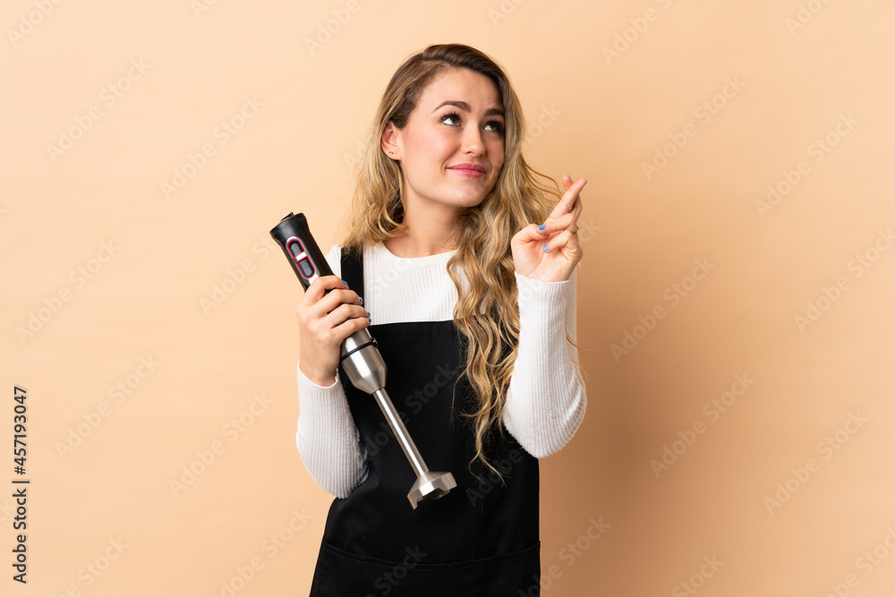 Young brazilian woman using hand blender isolated on beige background with fingers crossing and wishing the best Young brazilian woman using hand blender isolated on beige background with fingers crossing and wishing the best