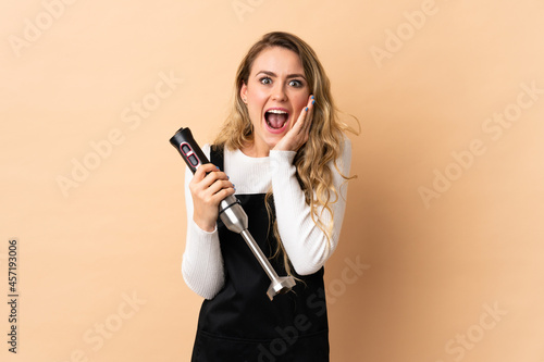 Young brazilian woman using hand blender isolated on beige background with surprise and shocked facial expression