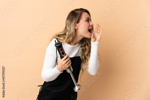 Young brazilian woman using hand blender isolated on beige background shouting with mouth wide open to the side
