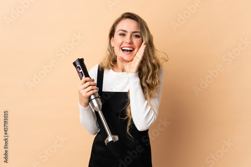 Young brazilian woman using hand blender isolated on beige background shouting with mouth wide open
