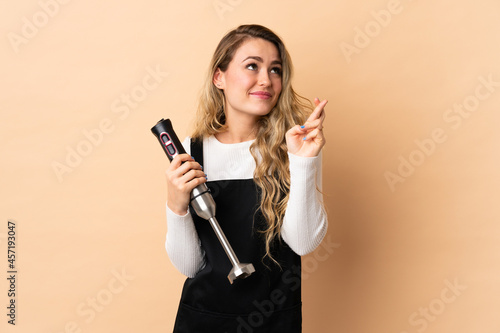 Young brazilian woman using hand blender isolated on beige background with fingers crossing and wishing the best