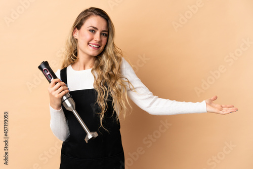 Young brazilian woman using hand blender isolated on beige background extending hands to the side for inviting to come