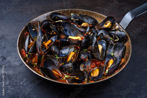 Traditional barbecue Italian blue mussel in tomato red wine sauce with herb and garlic as close-up in a rustic iron pan