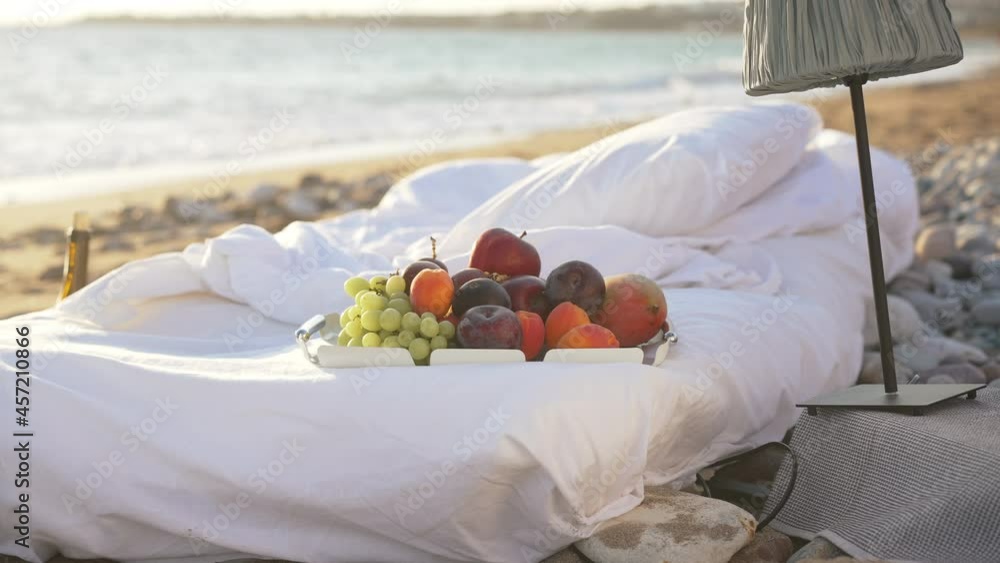 Exotic fruits on white soft comfortable bed on sandy beach in sunlight. Tasty vitamin grape peach plum and apple outdoors in sunshine with foamy blue Mediterranean sea waves rolling in slow motion Exotic fruits on white soft comfortable bed on sandy beach in sunlight. Tasty vitamin grape peach plum and apple outdoors in sunshine with foamy blue Mediterranean sea waves rolling in slow motion