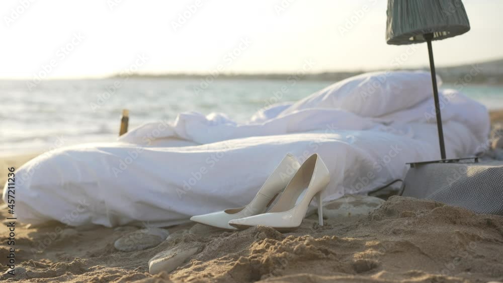 Wide shot white cozy soft bed on sandy beach with blue Mediterranean sea waves rolling at background in slow motion. Comfortable cushioned mattress and high-heels in sunshine outdoors on ocean coast Wide shot white cozy soft bed on sandy beach with blue Mediterranean sea waves rolling at background in slow motion. Comfortable cushioned mattress and high-heels in sunshine outdoors on ocean coast
