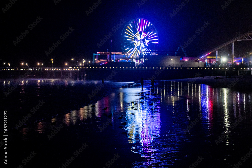 The lights of the Santa Monica Pier, the Ferris wheel and the Rollercoaster reflected in the water of the Pacific Ocean on Santa Monica Beach.