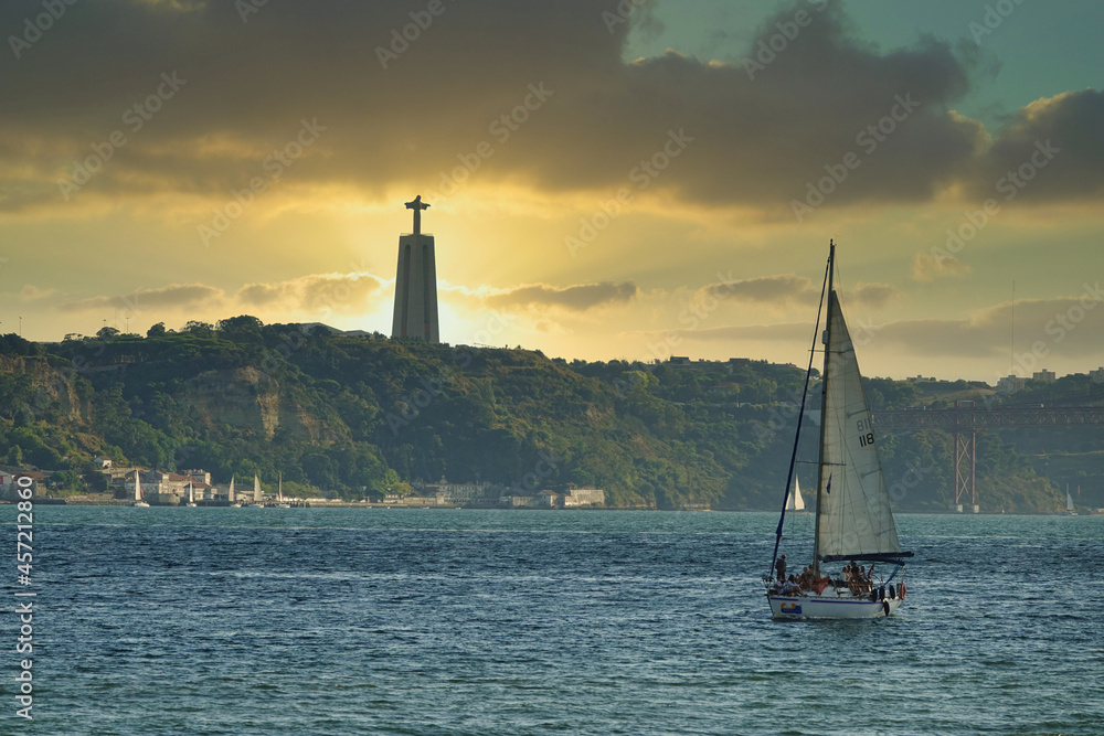 Sailboat sailing in Lisbon (Portugal) with the sunset sun behind the statue of Cristo Rei on the horizon