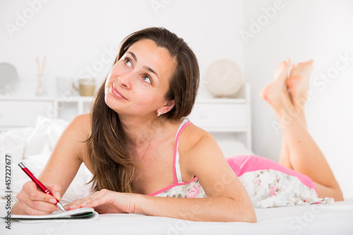 Portrait of young smiling woman lounging in bed and writing to diary in home interior photo