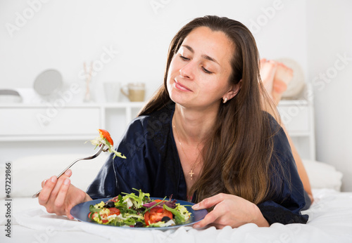 Young pretty woman eating vegetable salad from plato in bed at home