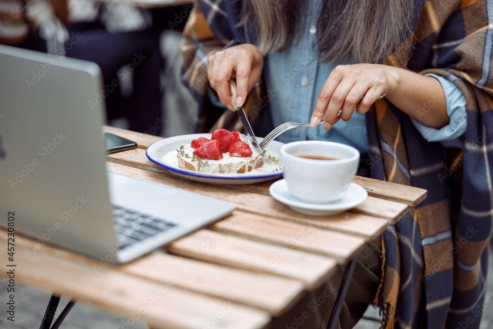 Mature woman eats tasty toast with cut strawberries and cream near laptop and cup of coffee at table on outdoors cafe terrace closeup