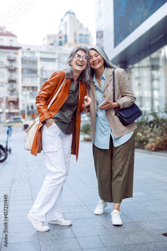 Stylish happy senior women companions stand on modern city street on nice autumn day. Friends spend time together