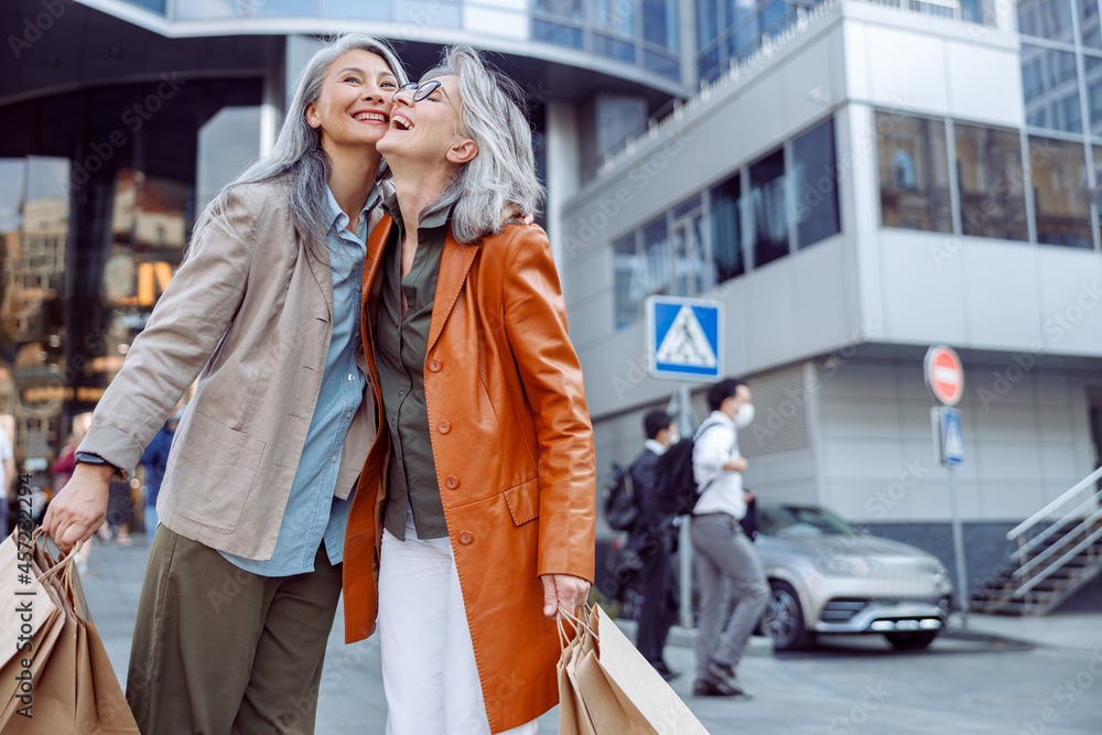 Fototapeta premium Joyful senior woman with paper shopping bags hug Asian lady on modern city street on autumn day. Friends spend time together