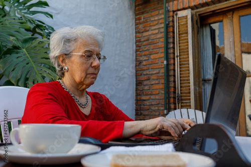 Older adult woman, working or studying on laptop while having breakfast at home.