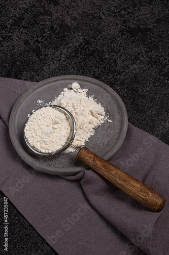 A strainer with a wooden handle with flour in a concrete plate on a black background. Ears of wheat and a linen napkin