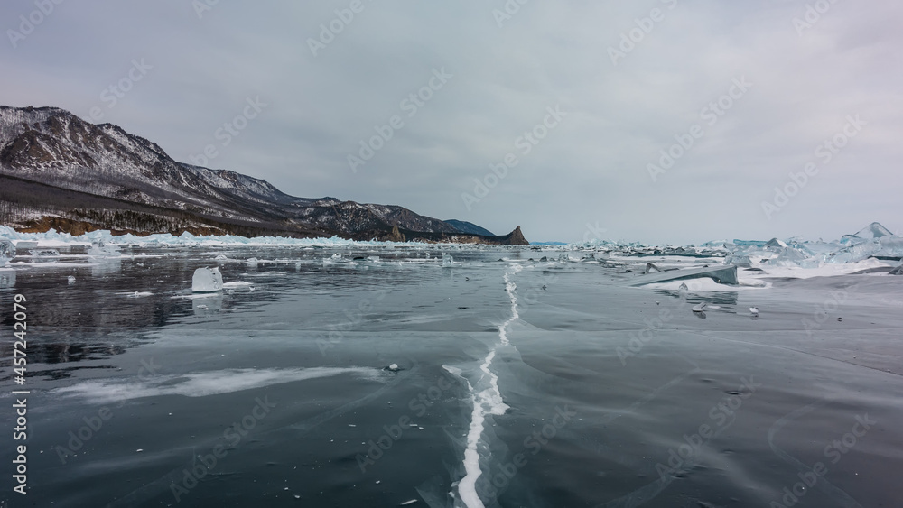 Obraz premium A deep crack is visible on the smooth surface of the frozen lake, fragments of ice are scattered. A mountain range against a cloudy sky. Reflection. Baikal