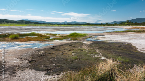 The river of hot springs flows in the caldera of an extinct volcano. The grass grows on stony soil. Bear paw prints are visible on the moss. A mountain range against the blue sky. Kamchatka. Uzon