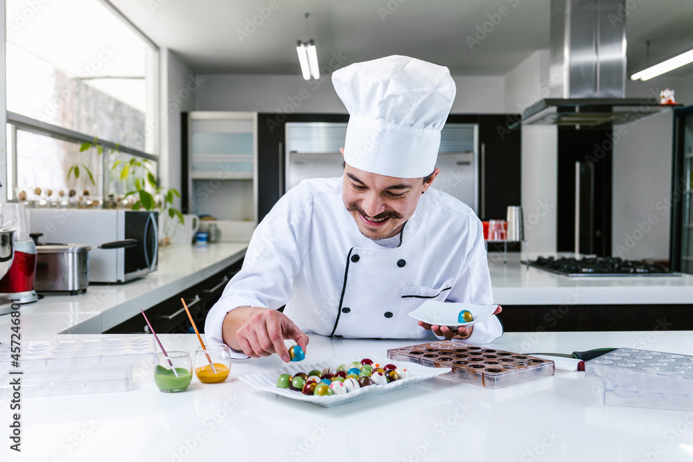 young mexican man chocolatier in chef hat standing with chocolates candies on plate in a commercial kitchen in Mexico Latin America young mexican man chocolatier in chef hat standing with chocolates candies on plate in a commercial kitchen in Mexico Latin America