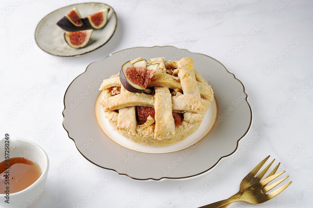 A sweet fig tart pastry on a sophisticated plate, two golden dessert spoons, honey, cut figs on a marble surface