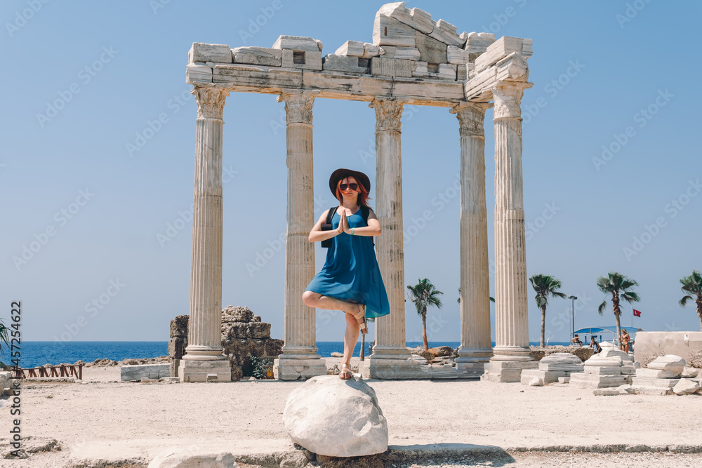Naklejka premium A beautiful girl in a dress, hat and sunglasses poses with a view of the ruins of the ancient Roman temple of Apolon. Side, Turkey.