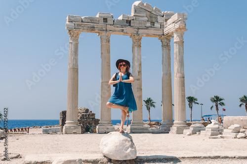 A beautiful girl in a dress, hat and sunglasses poses with a view of the ruins of the ancient Roman temple of Apolon. Side, Turkey.