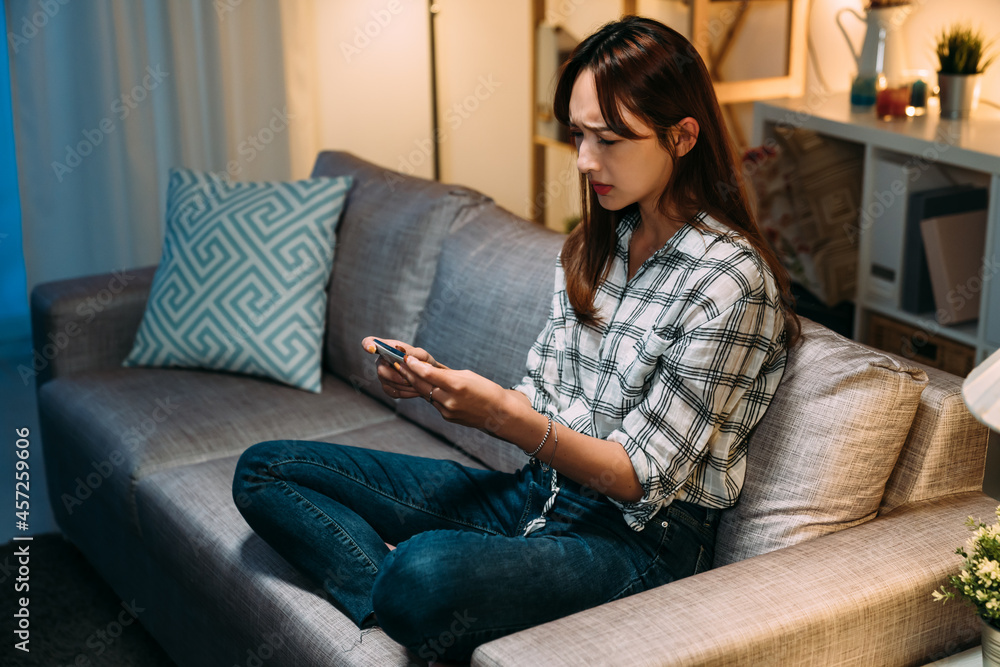 Obraz premium upset asian young woman playing mobile games is frowning at the phone in hands feeling unhappy about losing on the sofa in the living room at home.