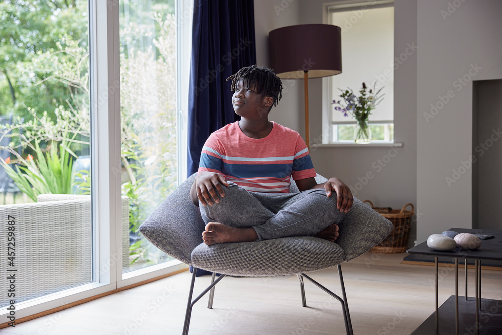 Thoughtful Teenage Boy Sitting Cross Legged On Chair At Home Looking Out Of Window Thoughtful Teenage Boy Sitting Cross Legged On Chair At Home Looking Out Of Window